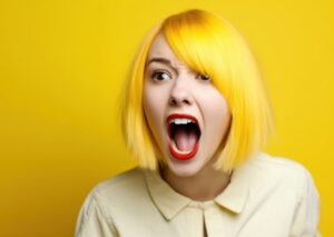 Kobieta z żółtymi włosami i czerwoną szminką, krzycząca z szeroko otwartymi ustami na tle żółtej ściany.